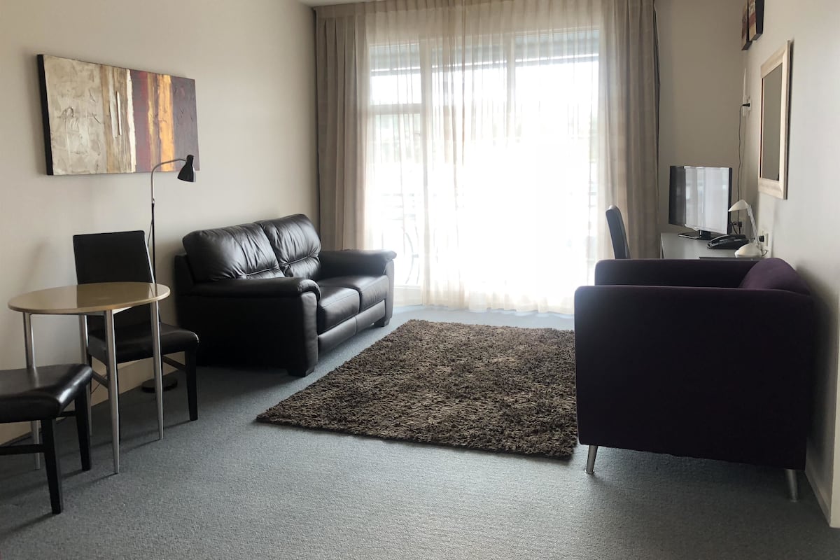 A living room with a black leather sofa, a small round table with two chairs, a dark rug, and a television on a stand.