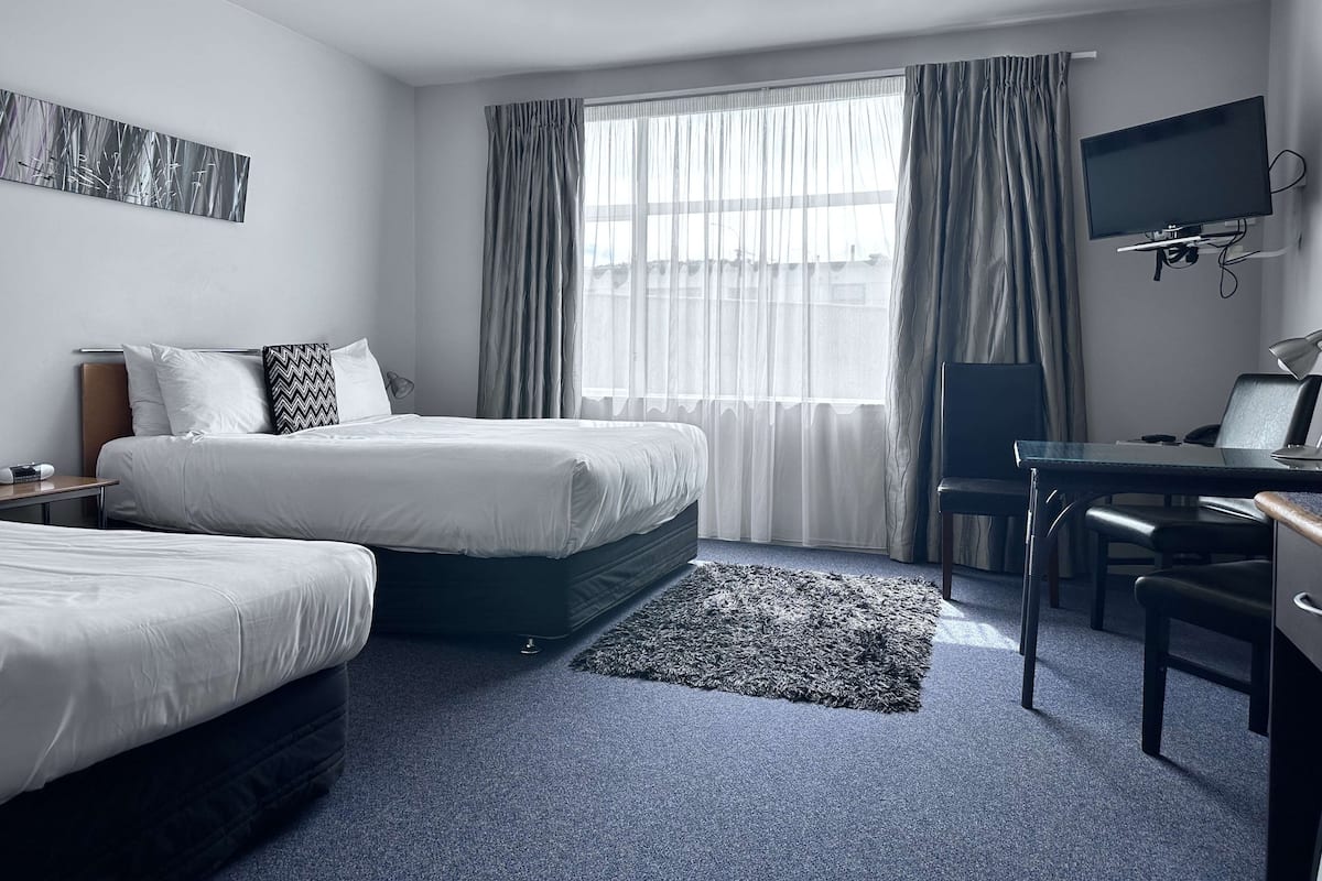 A hotel room with two beds, a desk with a chair, a television, and a large window with curtains.