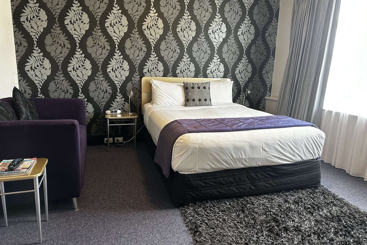 A hotel room with a bed, a small table, a chair, and a patterned wallpaper.