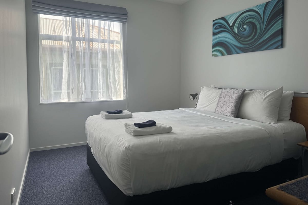 A hotel room with a large bed, a window with sheer curtains, a painting on the wall, and a small table with a lamp.