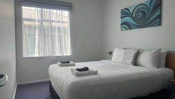 Habitación de hotel con una cama grande, una ventana con cortinas traslúcidas, un cuadro en la pared y una mesita con una lámpara.
