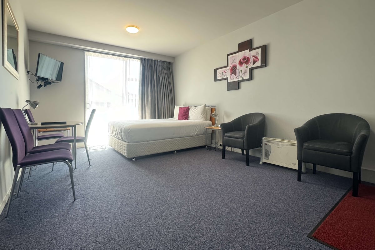 A hotel room with a bed, two chairs, a desk with a computer, a TV, and a window with curtains.