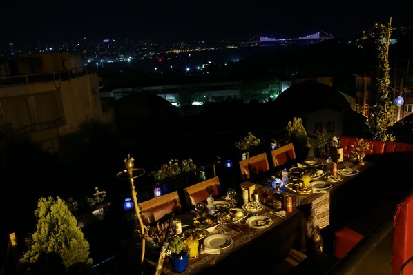 Un área de comedor en la azotea con una mesa dispuesta para una comida, con vista a un paisaje urbano nocturno.