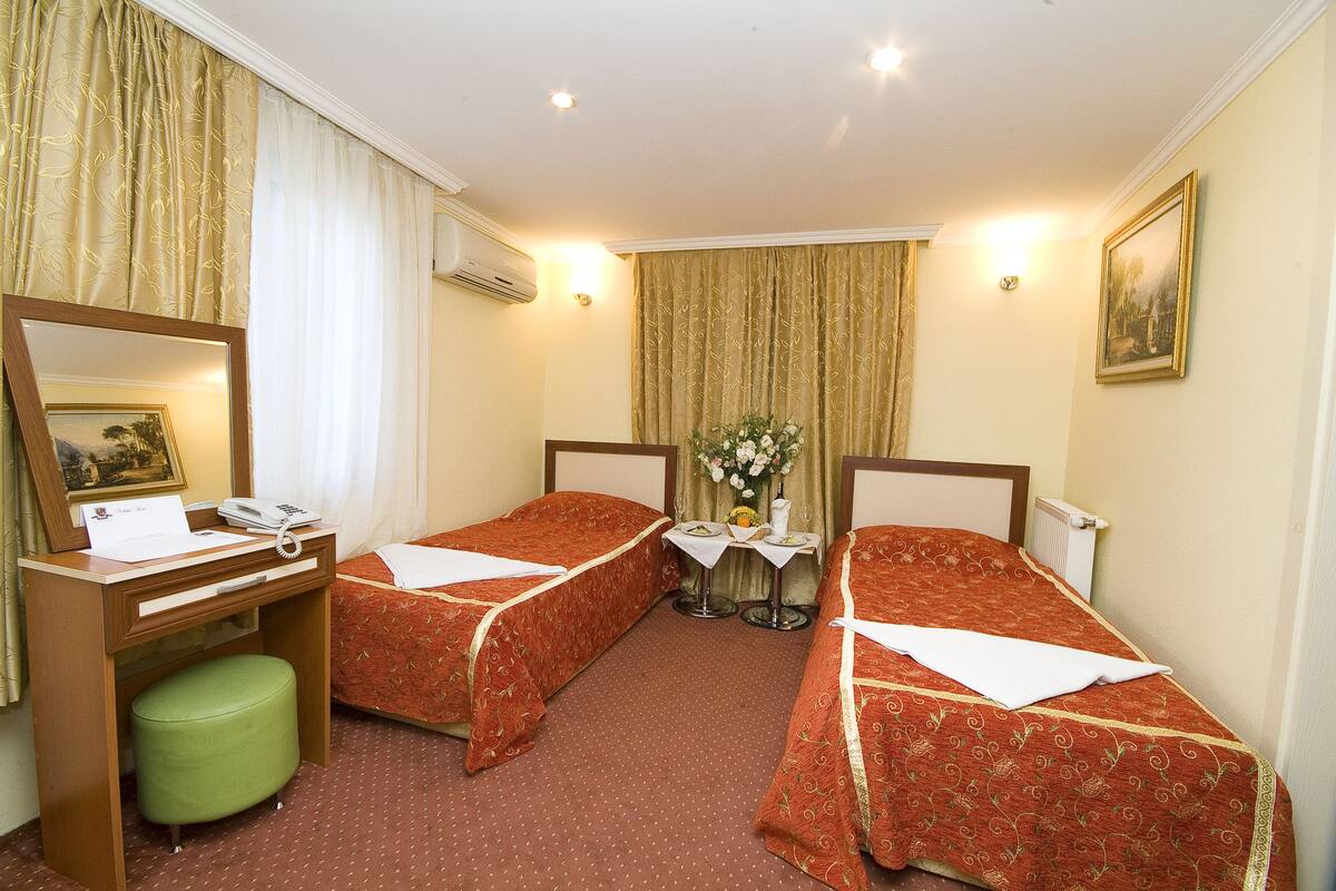 Quarto de hotel com duas camas de solteiro, uma escrivaninha com telefone, um banquinho verde, uma mesinha com um vaso de flores e um quadro pendurado na parede.