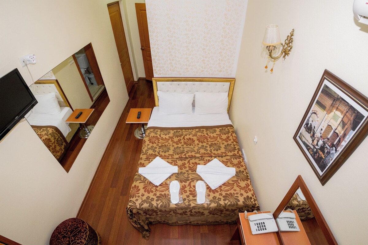 Habitación de hotel con cama, mesita de noche, espejo, lámpara de pared y un cuadro enmarcado.