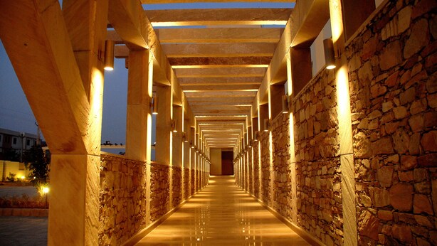 A corridor with stone walls and wooden beams, illuminated by overhead lights. - Cambay Grand (Amer)