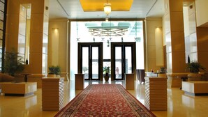 A spacious lobby with a red carpet leading to a reception desk, flanked by tall columns and potted plants. - Cambay Grand (Amer)