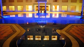 A large indoor swimming pool with a central fountain, surrounded by a tiled deck and a covered seating area. - Cambay Grand (Amer)