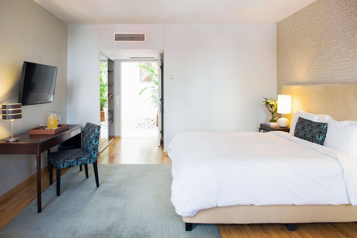 A hotel room with a bed, a desk with a chair, a television, and a nightstand with a lamp and a vase of flowers.