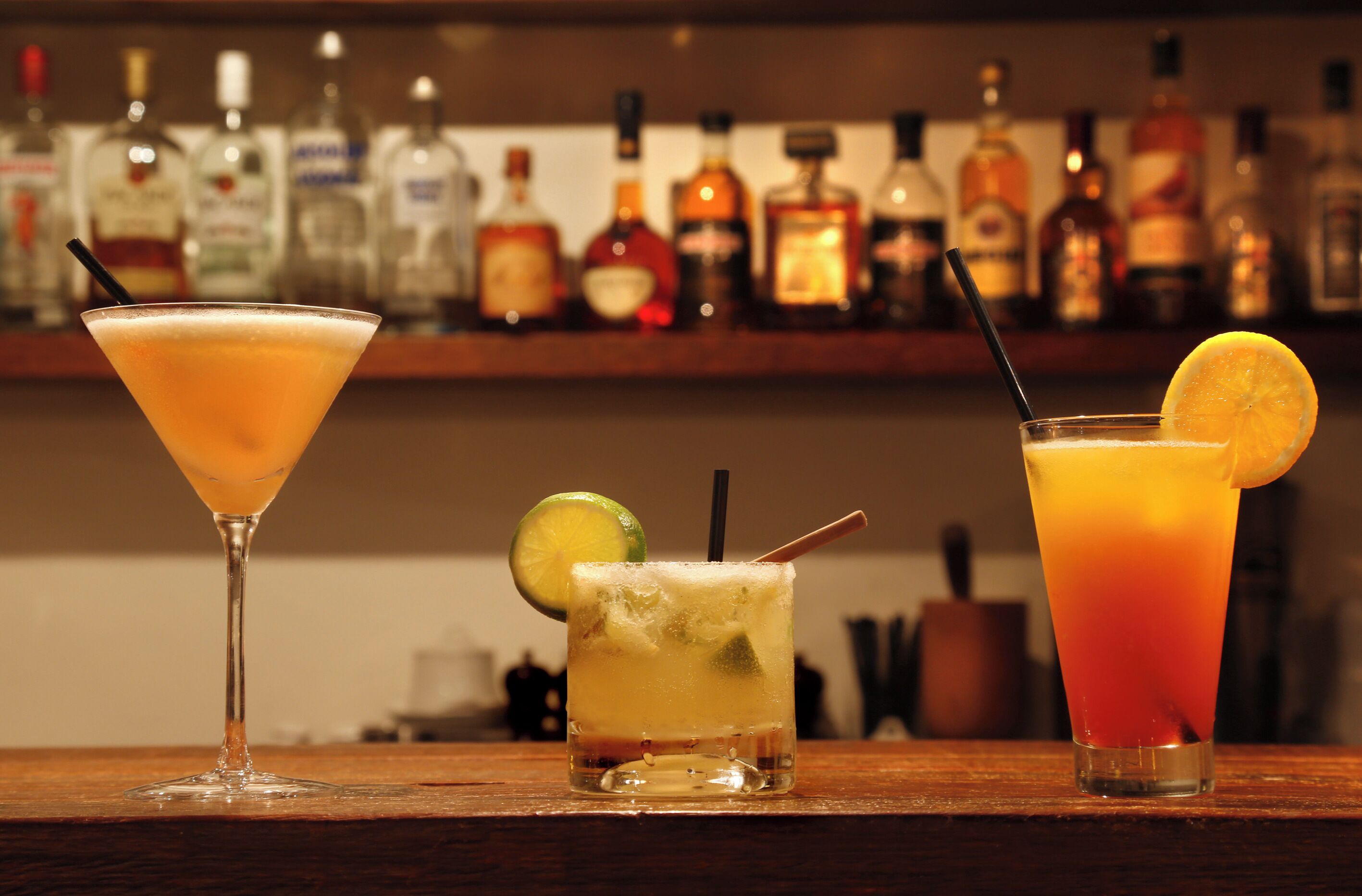 Drie verschillende cocktails op een bar met een verscheidenheid aan drankflessen op de achtergrond.