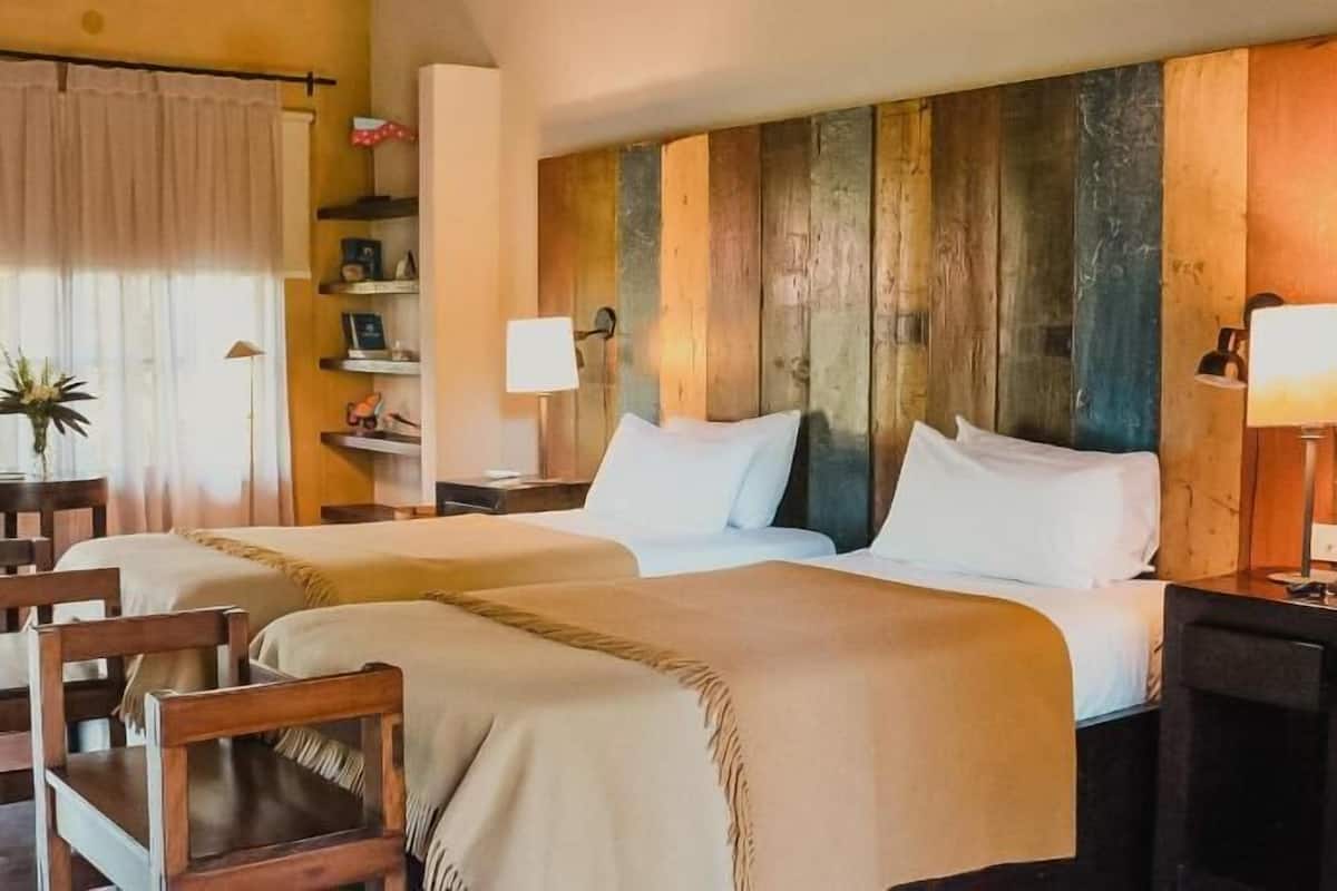 A hotel room with two beds, a desk, a chair, a lamp, and a shelf with various items.