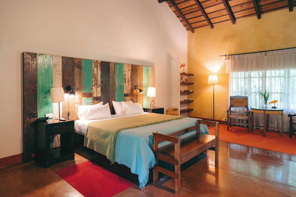A bedroom with a wooden headboard, a bed with white linens, a red rug, a wooden bench, a nightstand with a lamp, and a small table with a lamp.