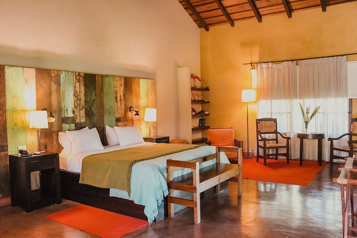 A hotel room with a bed, bedside tables, a chair, a small table, and a window with curtains.