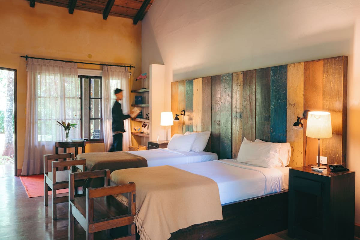 A hotel room with two beds, wooden headboards, a desk with a chair, and a window with curtains.