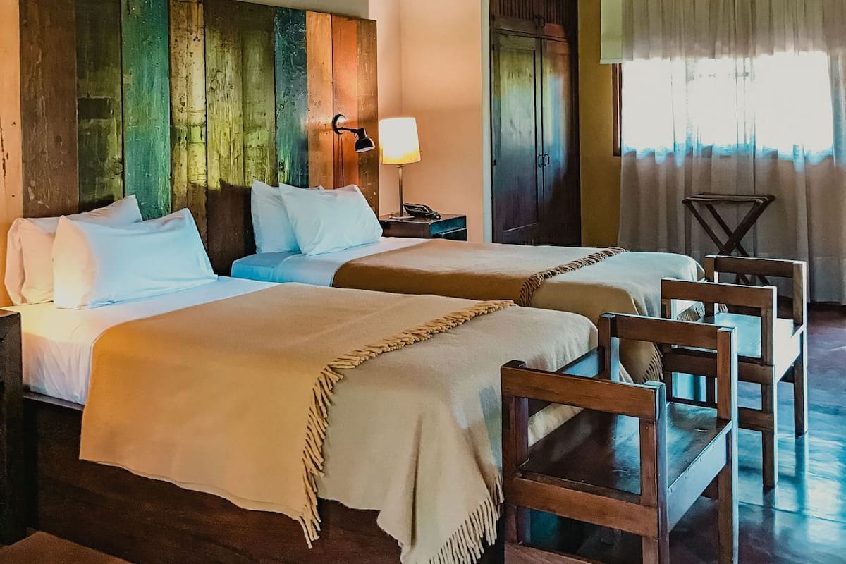 A bedroom with two beds, wooden headboards, a nightstand, a lamp, a wardrobe, and a window with curtains.