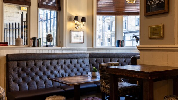 A cozy seating area with a cushioned bench, a wooden table, and a fireplace. - The Plough (Alnwick)