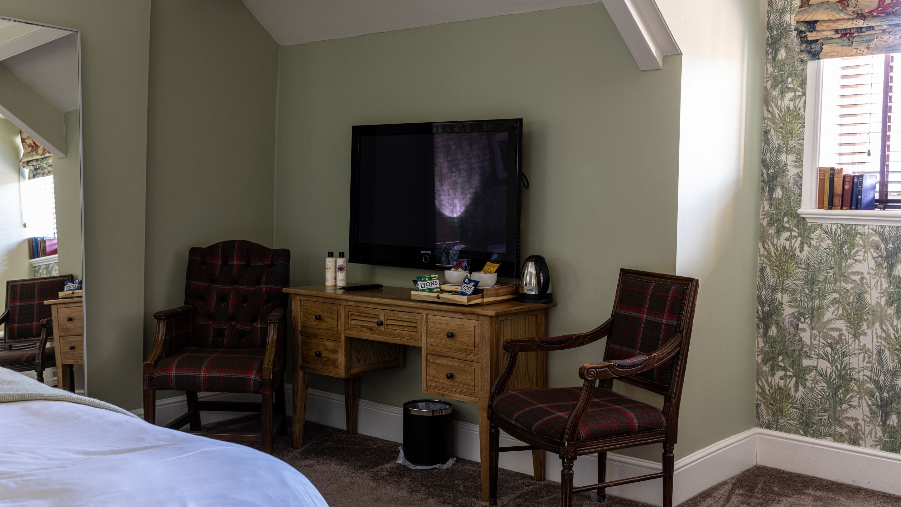 A bedroom with a bed, a desk with a television, two chairs, a small trash bin, and a window with books.