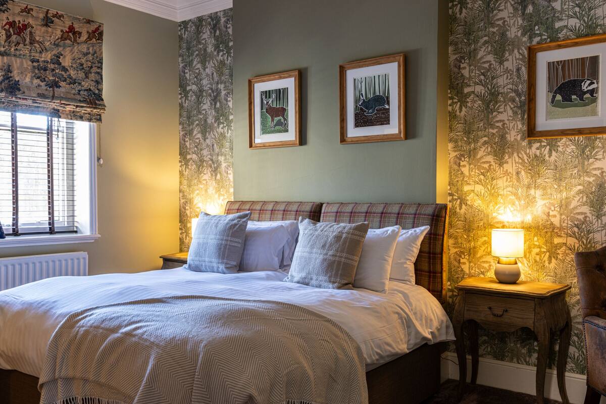 A bedroom with a bed, a window with blinds, a radiator, a nightstand with a lamp, and a chair.