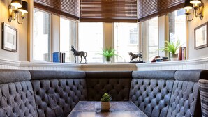 A corner seating area with cushioned benches, a table with a glass and a plant, and decorative items on the windowsill. - The Plough (Alnwick)