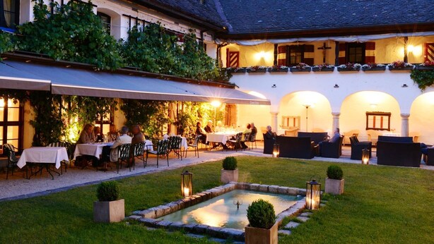 An outdoor dining area with tables and chairs, a small garden with a fountain, and a building with arches and a balcony. - Hotel Schloss Leonstain (Poertschach am Woerthersee)