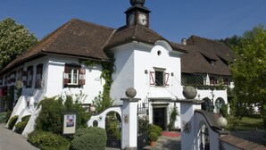 A white building with a clock tower, a red door, and a garden in front. - Hotel Schloss Leonstain (Poertschach am Woerthersee)