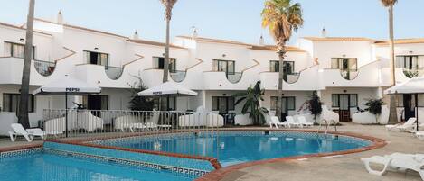 A swimming pool with lounge chairs and umbrellas, surrounded by a building with white walls and balconies.
