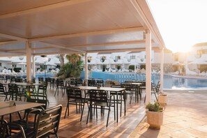 Un área de comedor al aire libre con mesas y sillas, una piscina y un edificio con balcones al fondo.