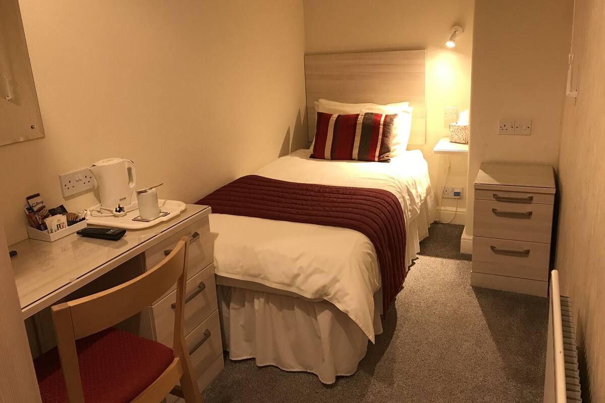 A compact hotel room with a bed, bedside table, desk with chair, and a small wardrobe.
