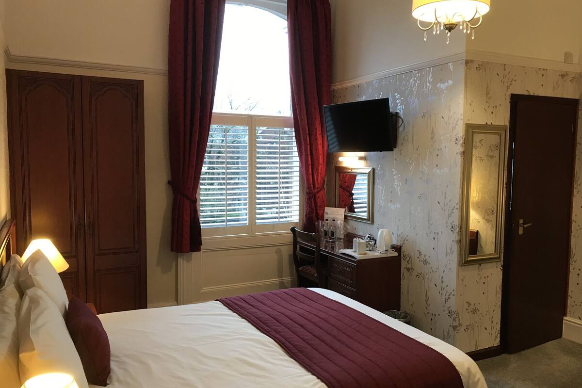 A hotel room with a large bed, a desk with a mirror, a flat-screen TV, and a window with curtains.