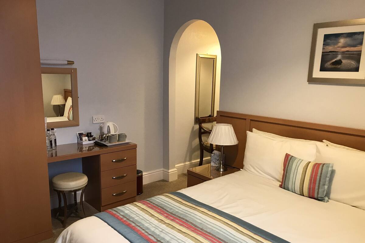 A hotel room with a bed, a desk with a chair, a lamp, and a mirror.