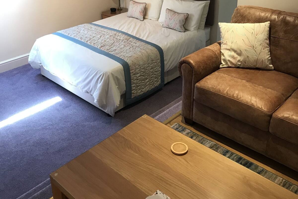 A bedroom with a bed, a brown leather sofa, a wooden coffee table, and a small table with a tissue box.