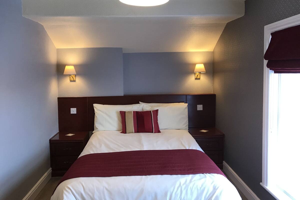 A hotel room with a bed, two bedside tables, a nightstand, a window with a blind, and two wall-mounted lamps.