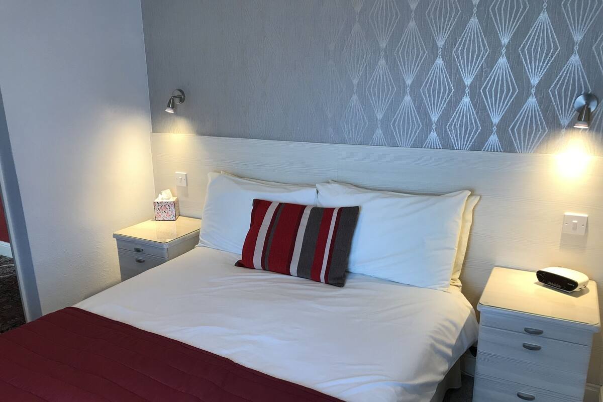 A neatly made bed with white linens and a red and gray striped pillow, flanked by bedside tables with lamps and a small bedside clock.