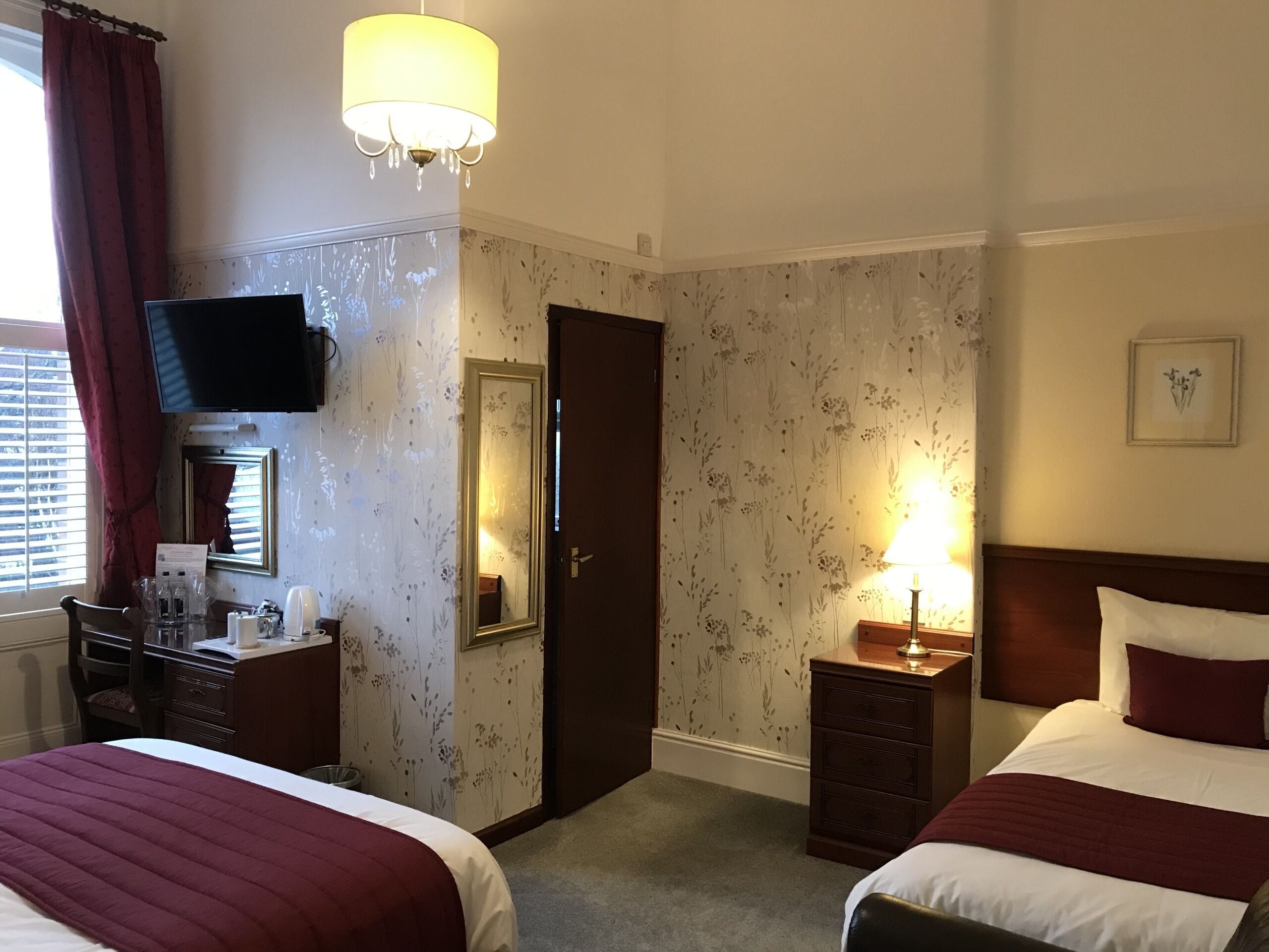 Ein Hotelzimmer mit zwei Betten, einem Schreibtisch mit Stuhl, einem Fernseher, einem Spiegel, einer Tür und einer Wand mit floralem Wandpapier.
