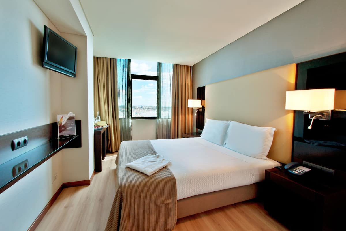 Ein Hotelzimmer mit einem großen Bett, einem an der Wand montierten Fernseher, einem Nachttisch mit Lampe und einem Fenster mit Vorhängen.