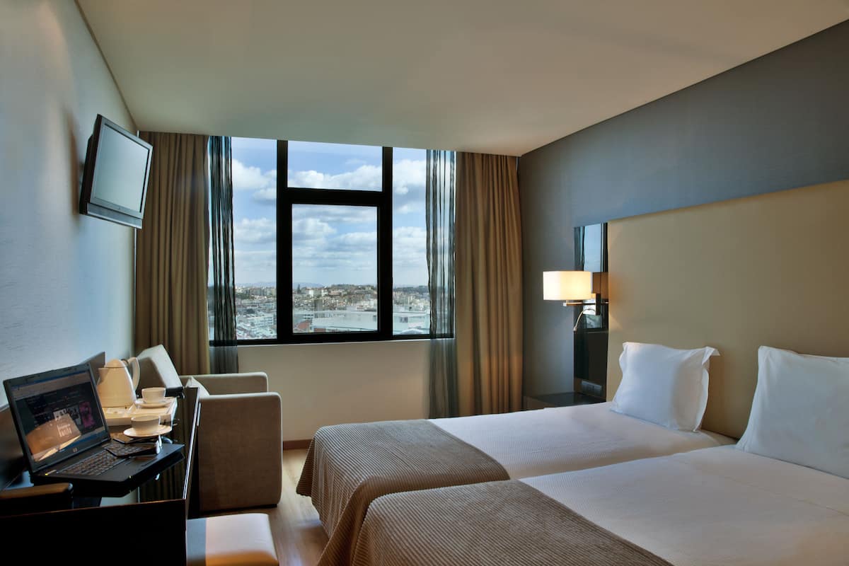Ein Hotelzimmer mit Bett, Schreibtisch mit Laptop, Stuhl, Fernseher und Fenster mit Blick auf die Stadt.