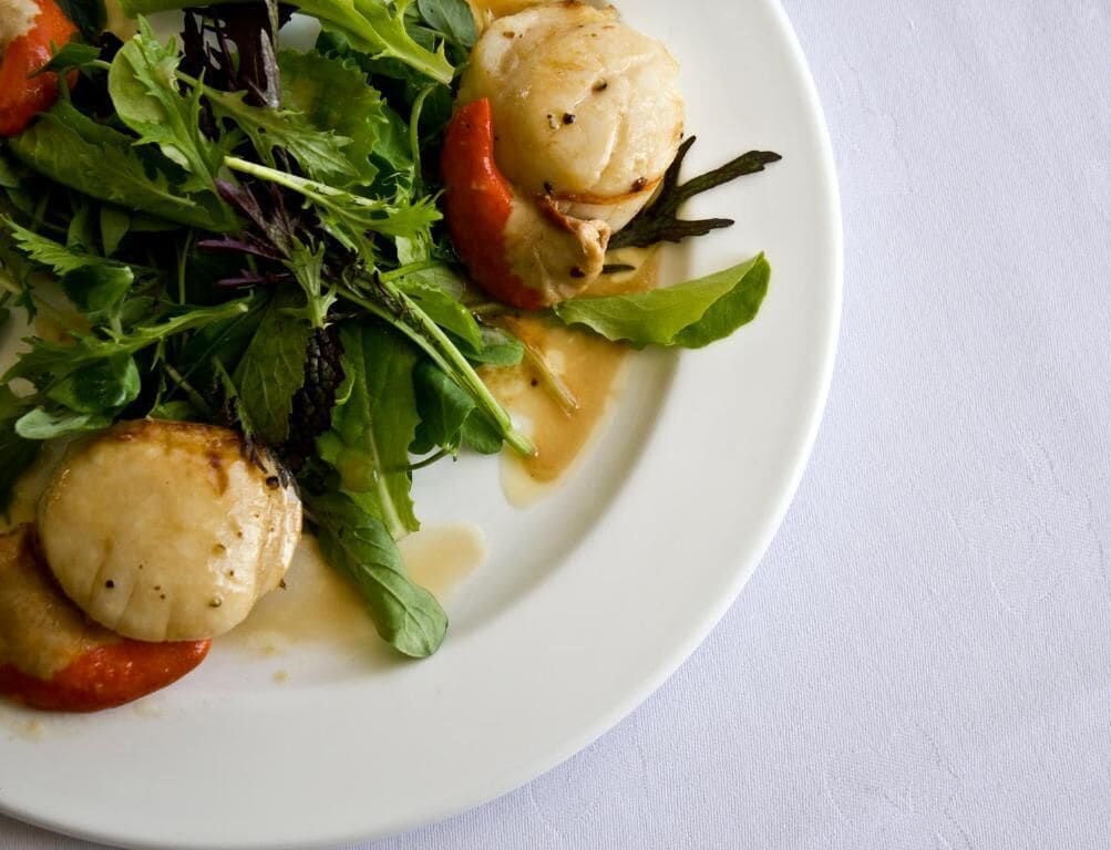Un plato con vieiras a la plancha, vegetales verdes y un pimiento rojo.
