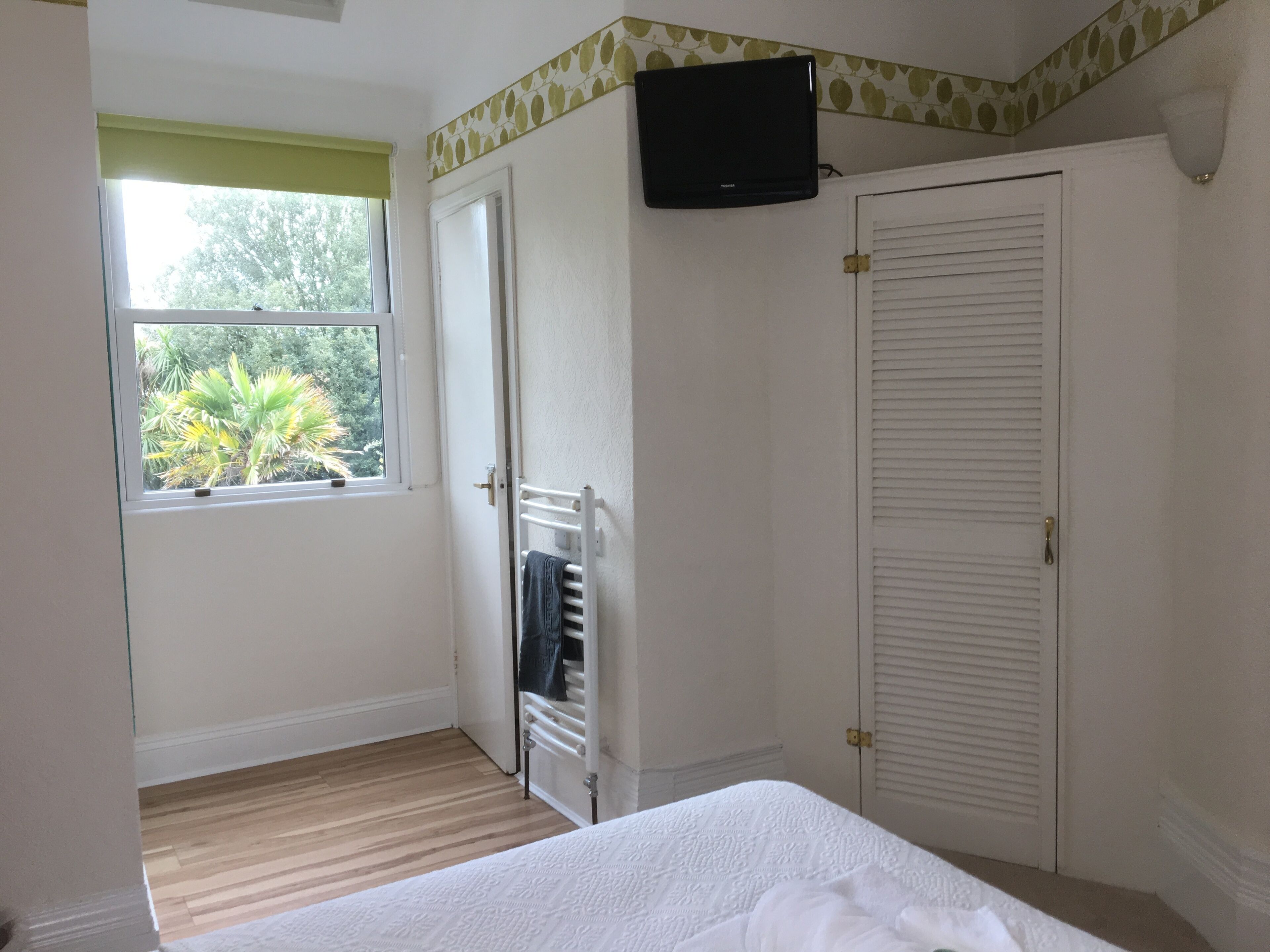A bedroom with a bed, a window with a view of greenery, a radiator, and a closed door.
