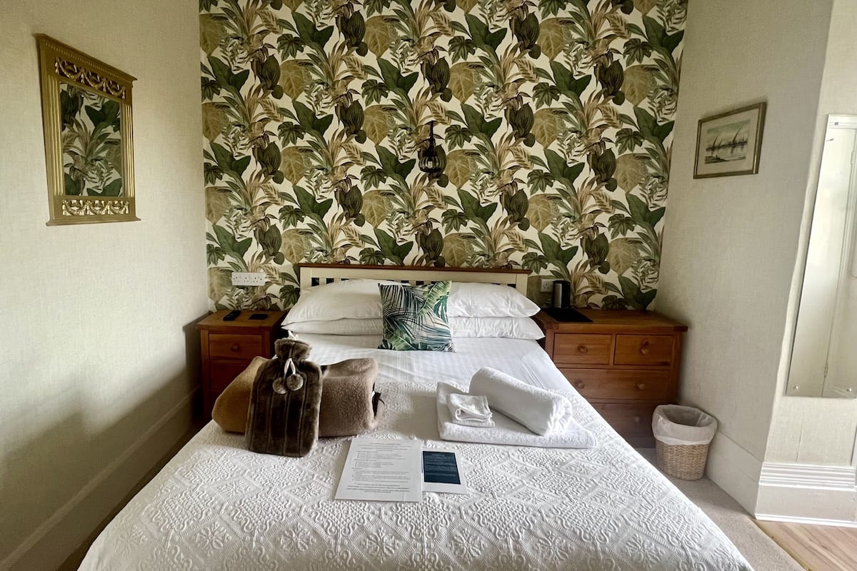 A neatly made bed with a floral wallpaper, a wooden nightstand, and a small trash bin.