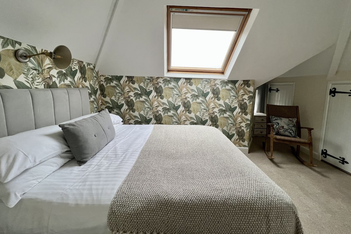 A bedroom with a sloped ceiling, a skylight, a bed with a grey headboard, a patterned wall, and a wooden chair.