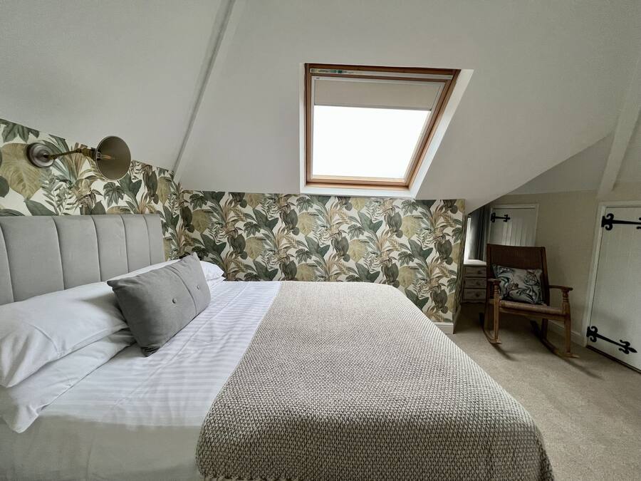 Quarto com teto inclinado, claraboia, cama com cabeceira cinzenta, parede estampada e cadeira de madeira.
