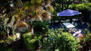 A patio area with a table and chairs under a blue umbrella, surrounded by lush greenery and various plants. - The Charterhouse (Torquay)