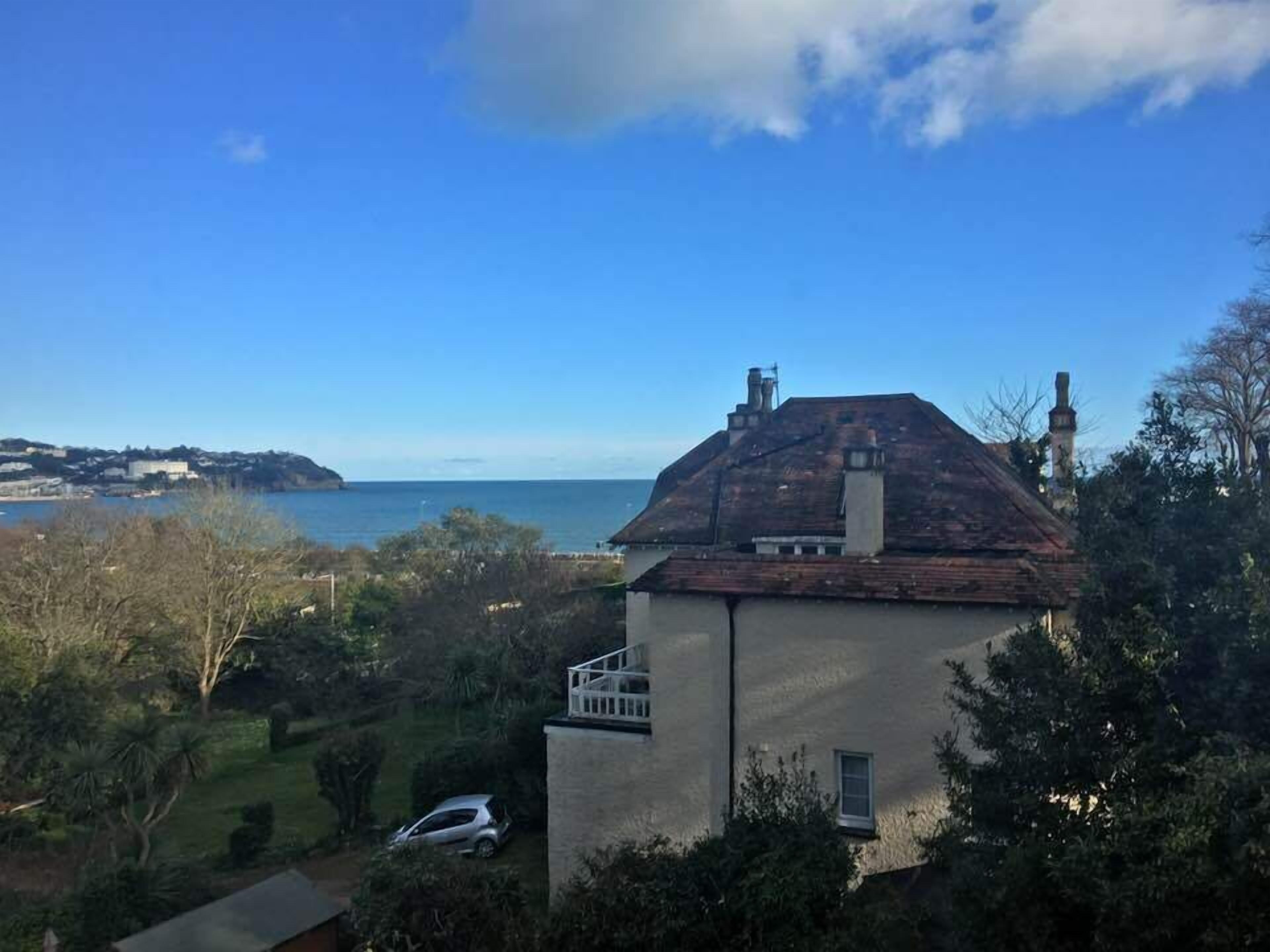 A house with a view of the sea, surrounded by greenery and a parked car.