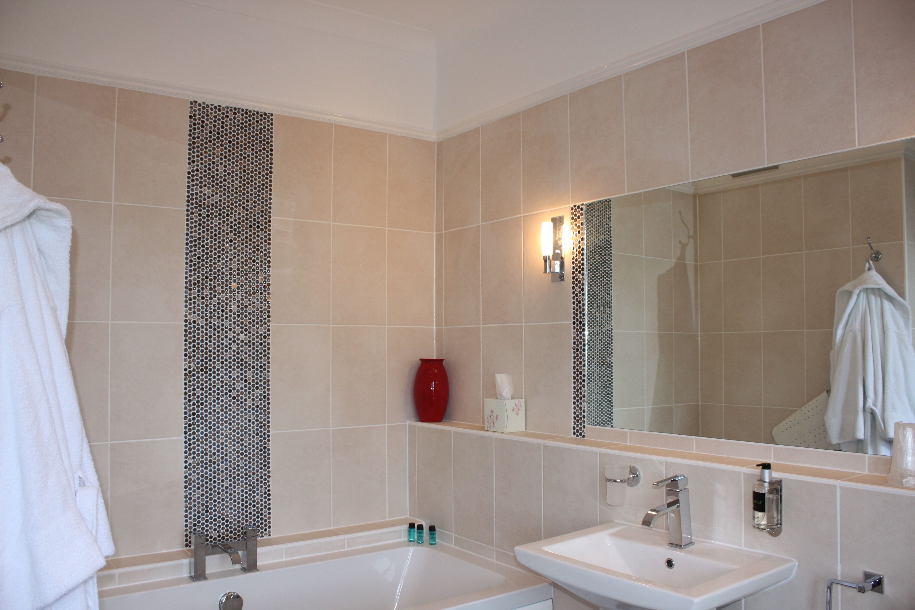 A bathroom with a bathtub, a sink, and a large mirror. There are towels hanging on the wall and a red vase on the shelf.