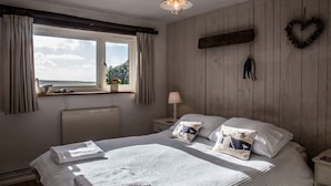 A bedroom with a large bed, a window with curtains, a wall-mounted light fixture, and a heart-shaped decoration. - Cheristow Farm Cottages (Bideford)
