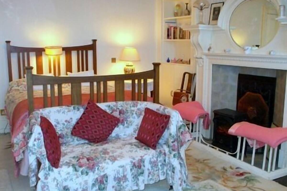 A cozy bedroom with a floral-patterned bed, a wooden chair, a small table, and a fireplace.