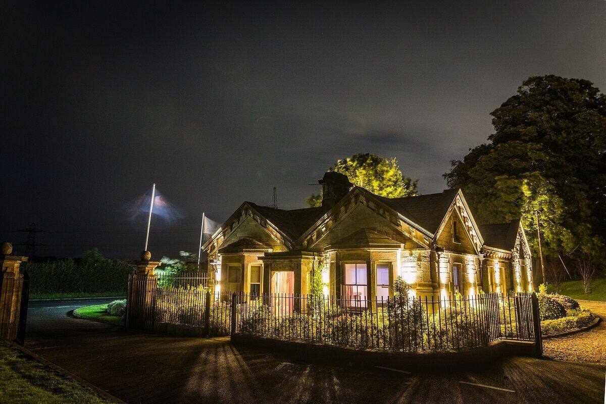 A well-lit, two-story building with a traditional architectural style, featuring a gated entrance and a driveway leading to the main entrance.