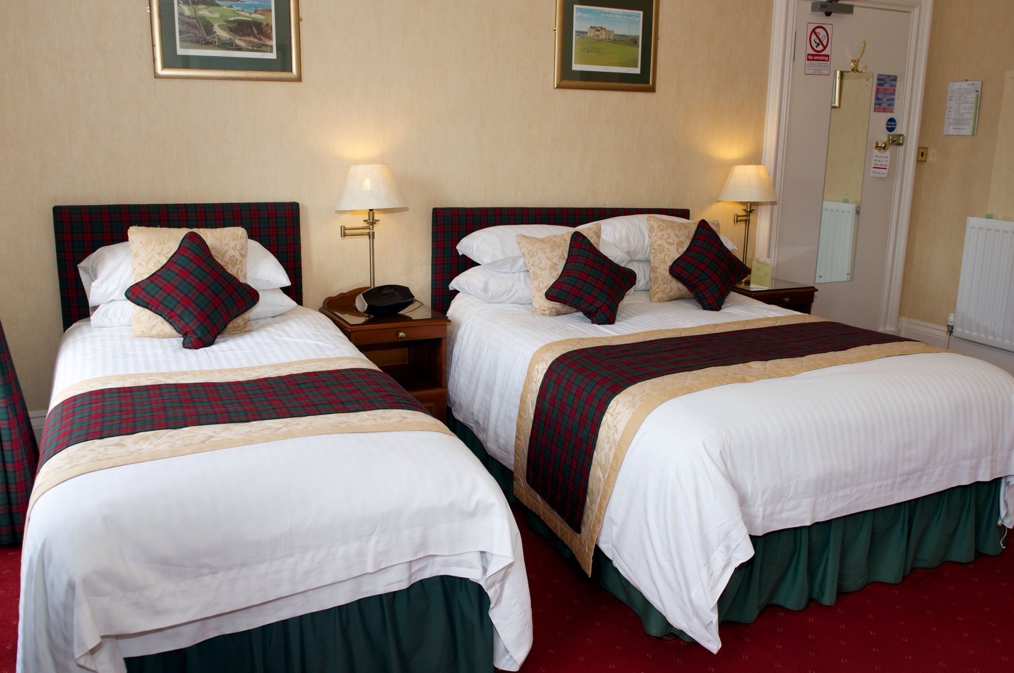A hotel room with two beds, each with a plaid bedspread and matching pillows. There are bedside tables with lamps, a door with a sign, and a wall-mounted light fixture.