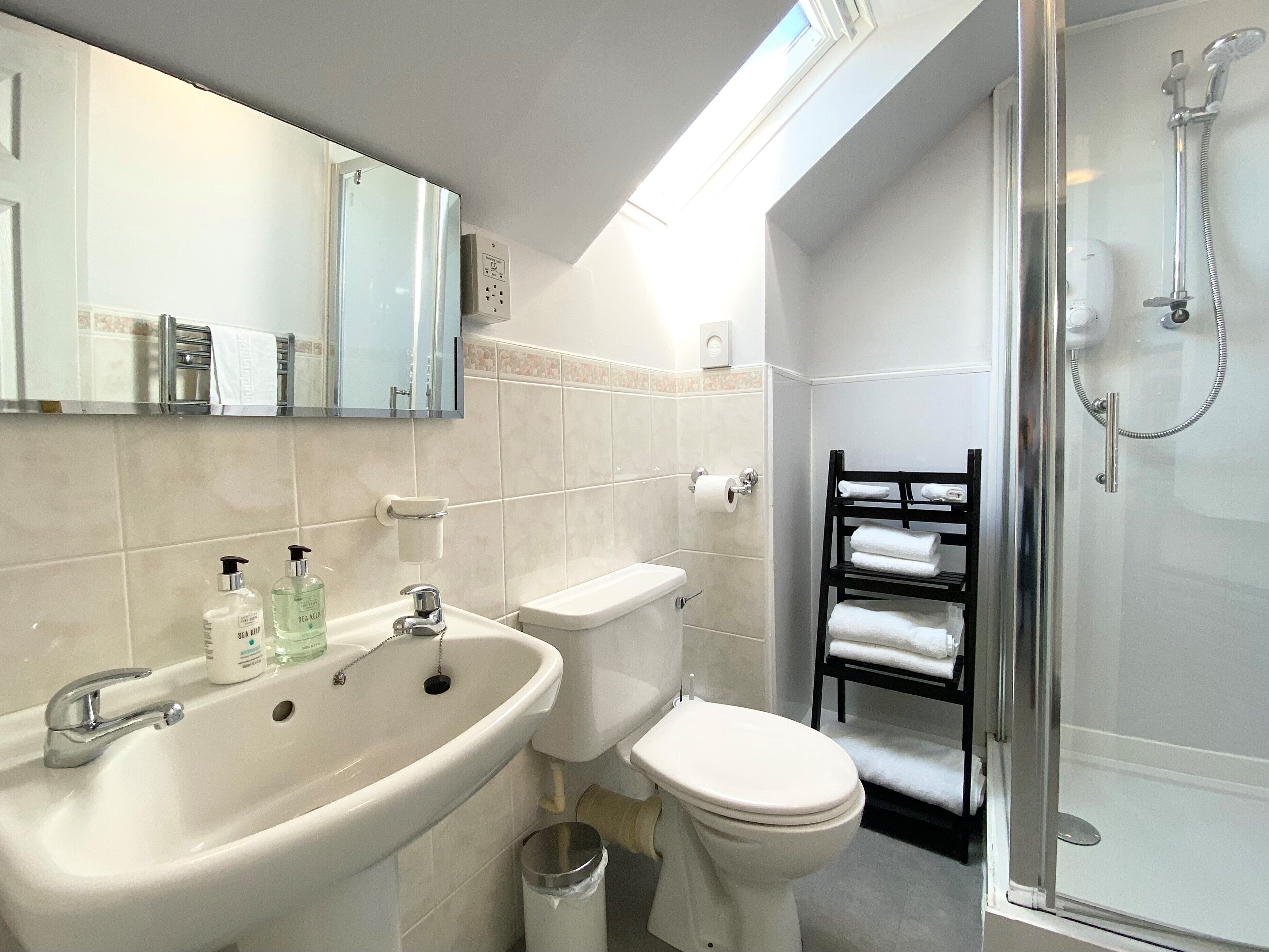A modern bathroom with a white sink, toilet, and shower. There is a towel rack with towels, a mirror, and a window above the sink.