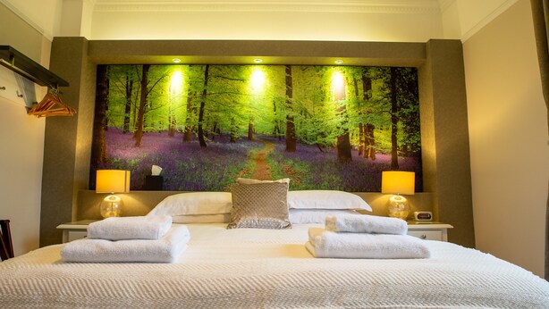 A hotel room with a large forest-themed mural on the wall, a neatly made bed with two towels, two bedside lamps, and a nightstand with a lamp and a clock. - Deanfield Bed and Breakfast (Lydney)